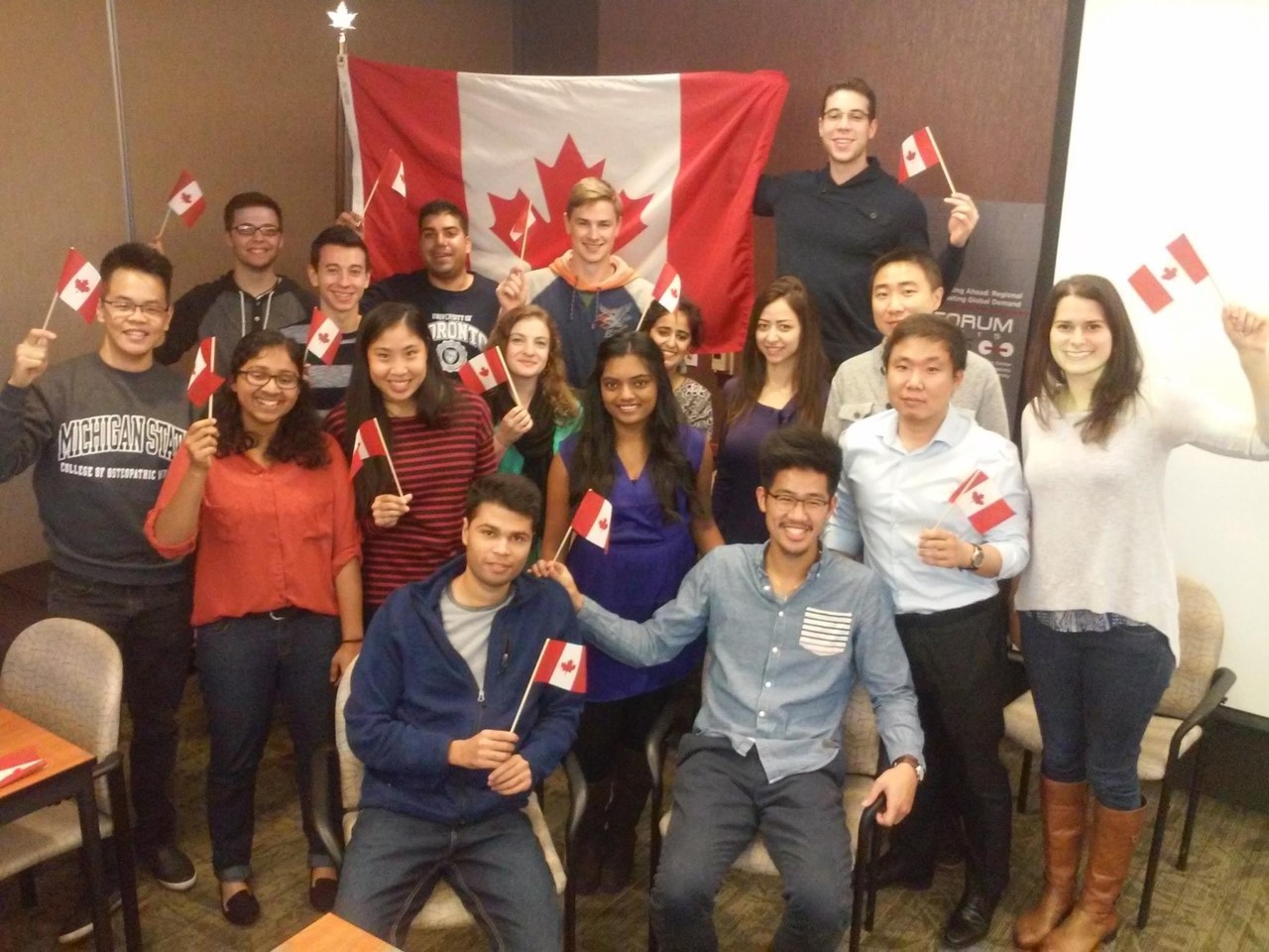 A diverse group of smiling students, standing in front of the Canadian flag. Some are holding small Canadian flags.